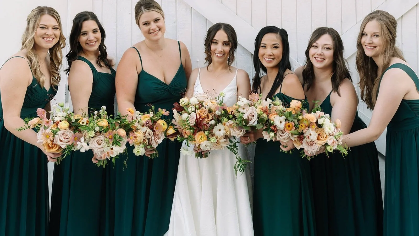 The bride and bridesmaids in deep green gowns hold whimsical bouquets with ranunculus, roses, and cosmos.