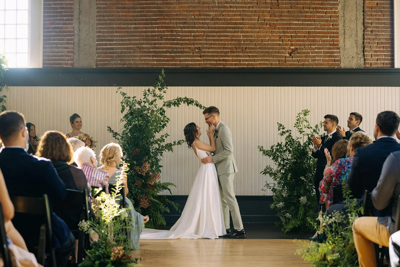 Bride and groom share their first kiss surrounded by lush greenery and golden sunlight at The Evergreen in Portland.