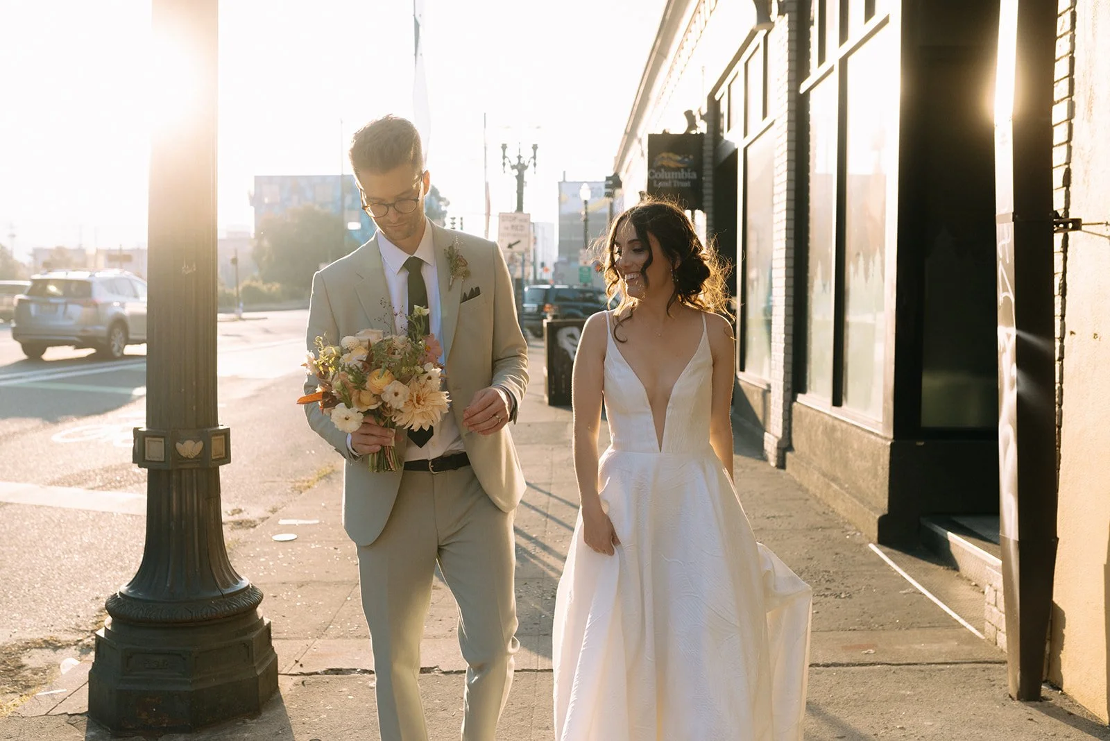 The bride and groom walk hand-in-hand through Portland’s golden hour glow with a lush bouquet of fall florals from Roots of Joy.