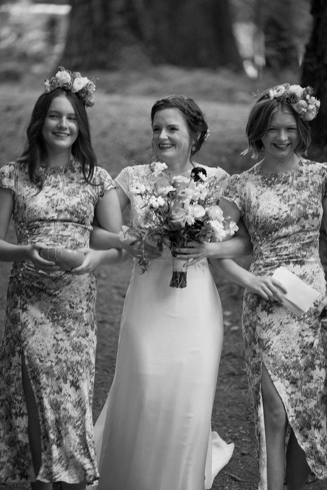 The bride smiles alongside bridesmaids wearing matching floral dresses and flower crowns, holding a garden-style bouquet filled with seasonal blooms.