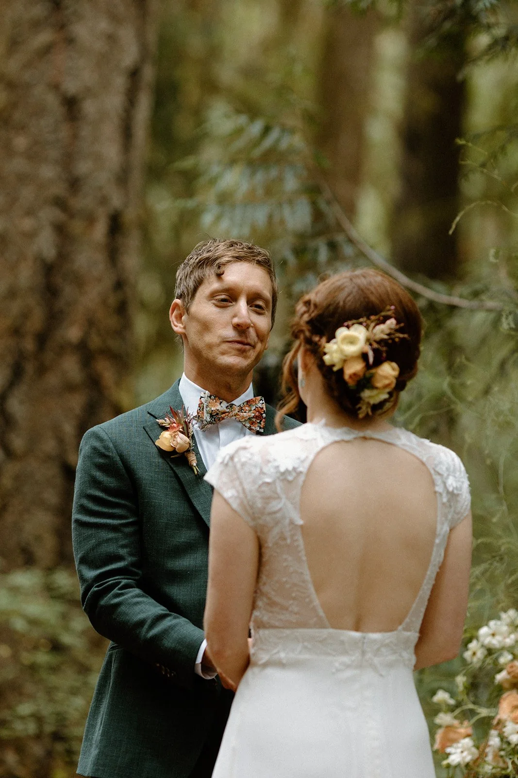 Bride and groom exchange vows beneath a rustic wooden arbor adorned with blush and cream florals at Camp Colton in the Oregon woods.