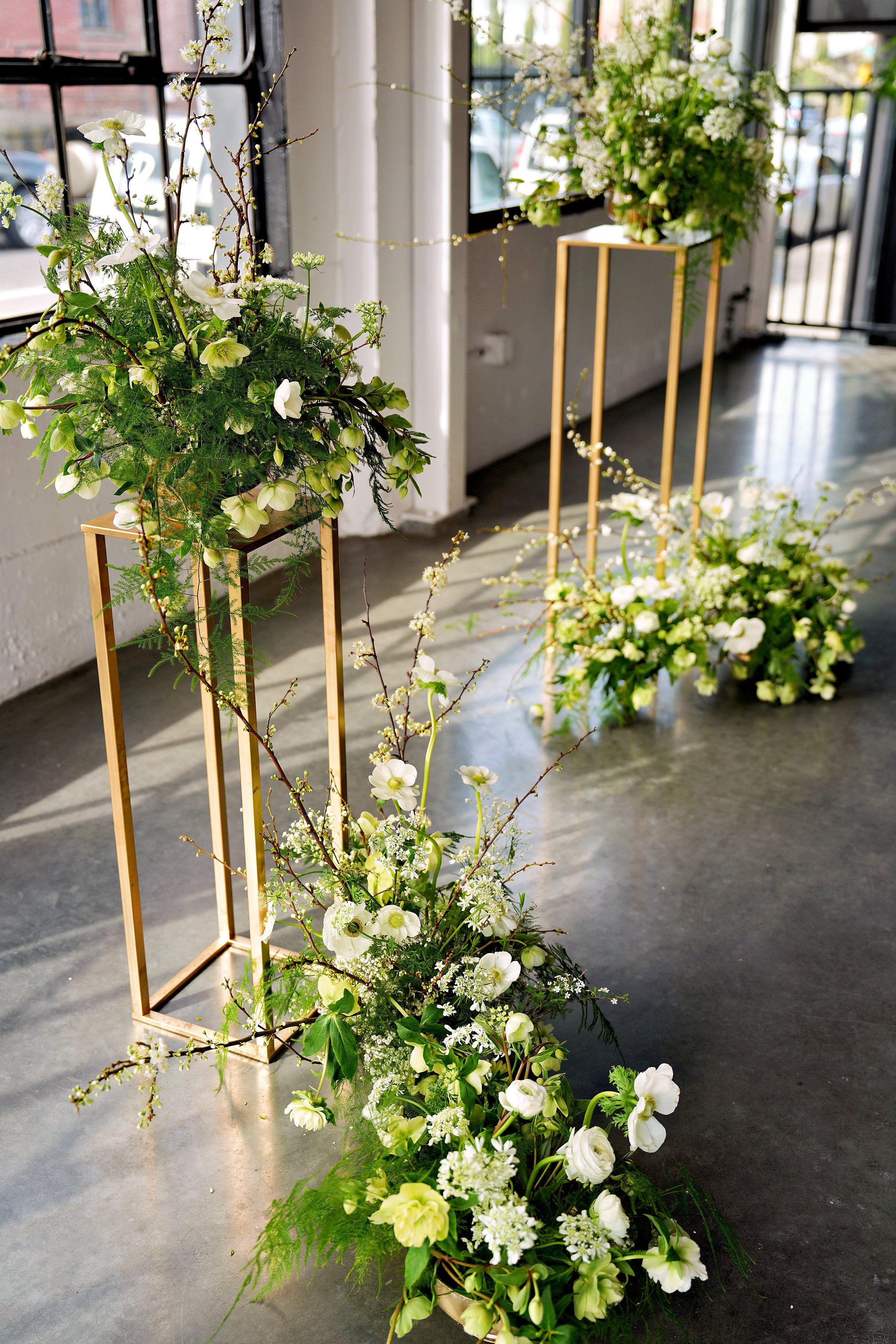A golden pedestal garden ceremony design featuring layered spring blooms in soft whites and greens, with climbing branches adding height and movement.