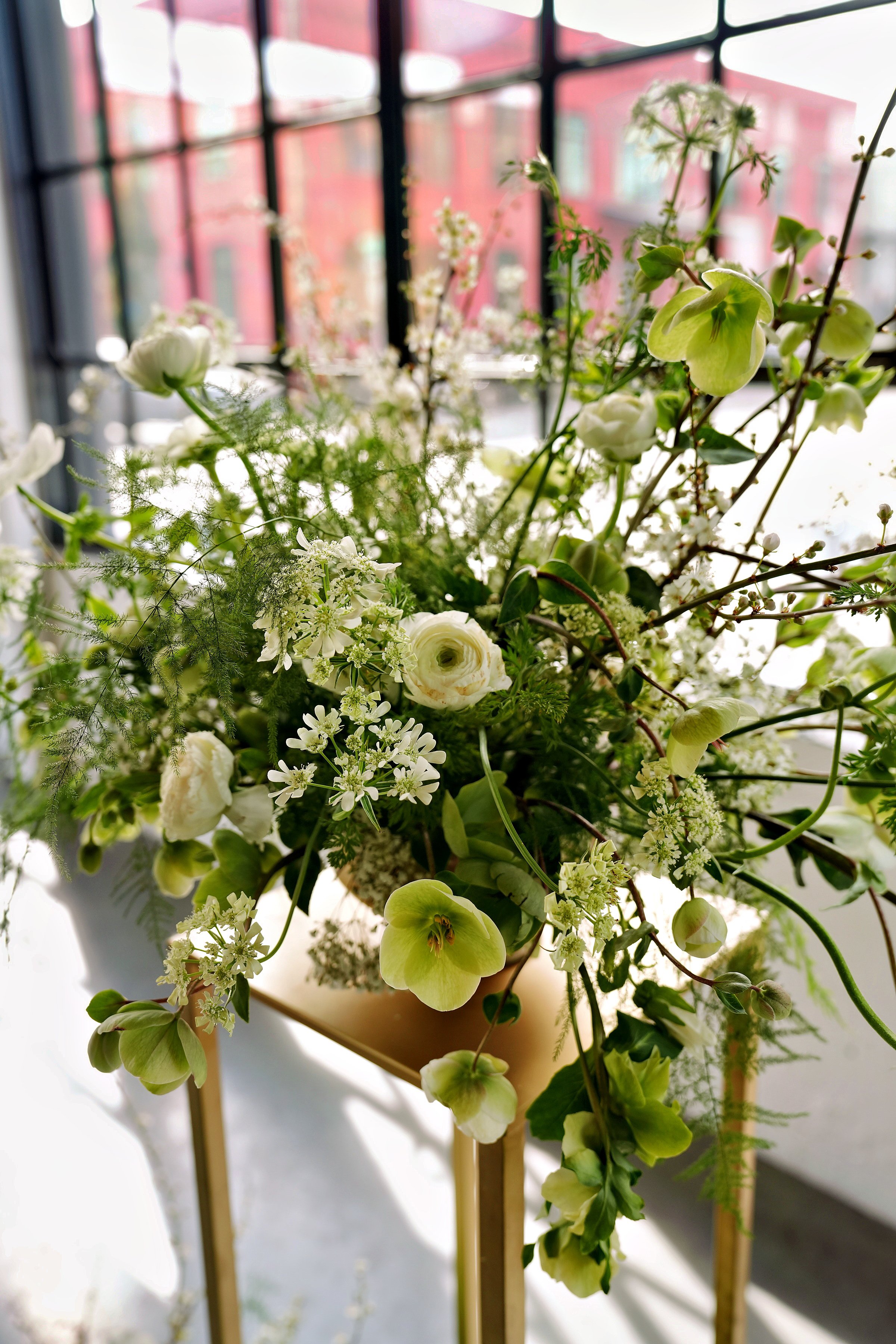 Modern pedestal floral arrangement of green hellebores, white ranunculus, and textural greenery displayed on a gold stand at an industrial wedding venue in Portland, OR.