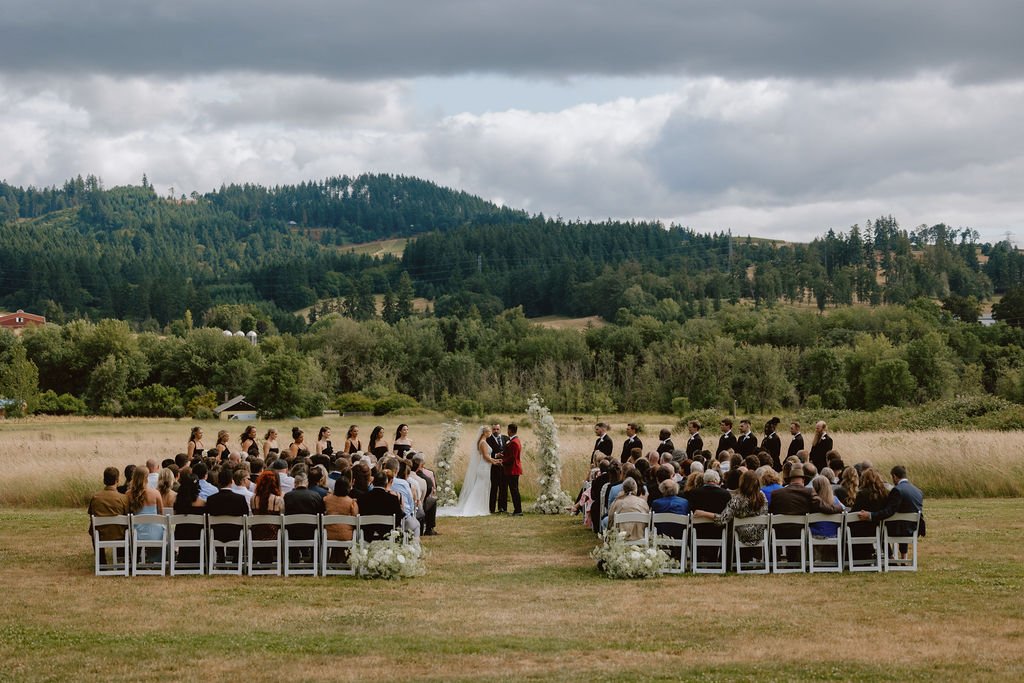 Set against rolling hills and the lush landscape of Oregon wine country, this wide-angle view captures the full magic of the ceremony. Rows of guests, a stunning wedding party, and delicate baby's breath pillars frame the couple’s vows under dramatic