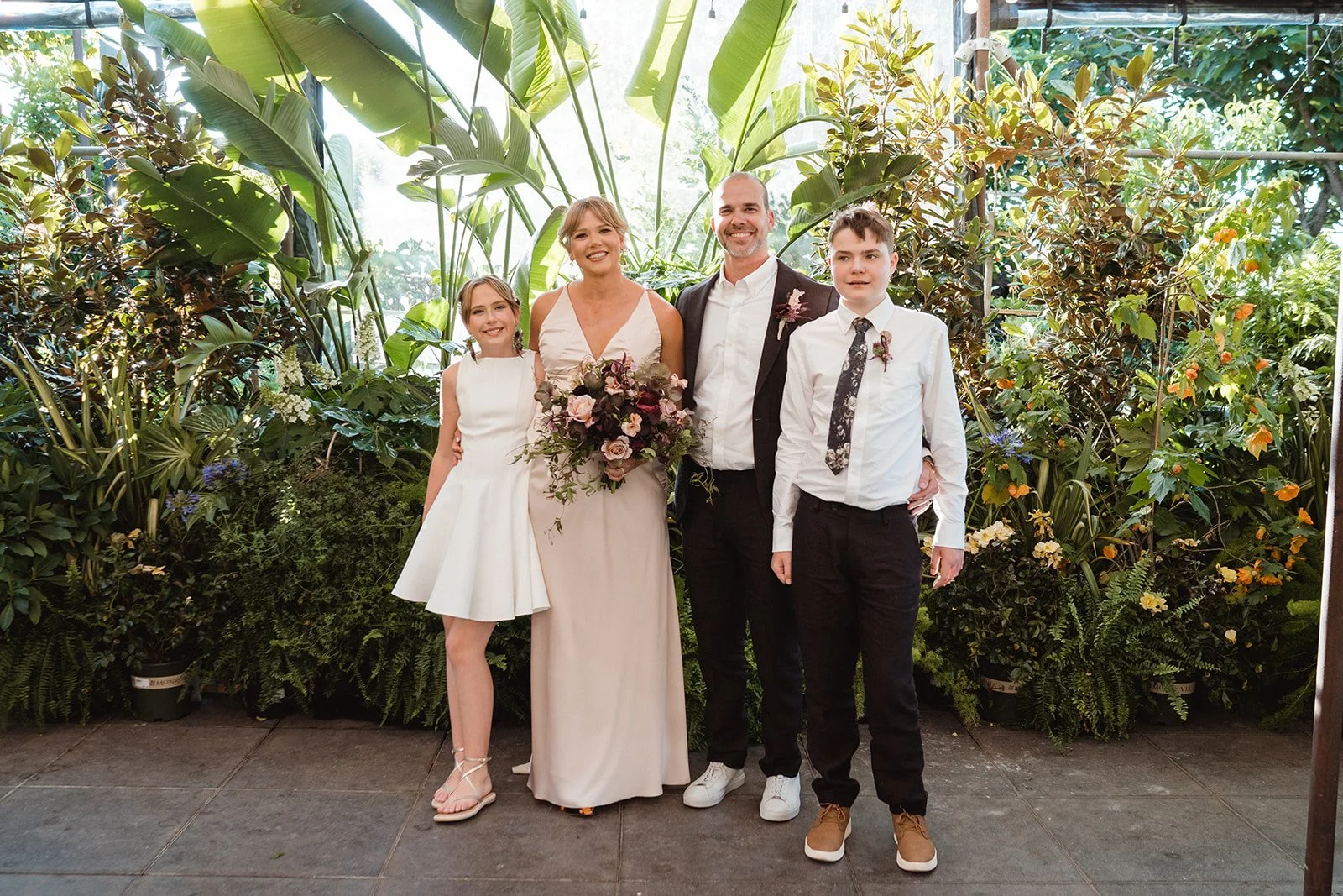 Bride, groom, and children pose in front of a lush plant wall, with a moody bouquet crafted by Roots of Joy for their Portland wedding.