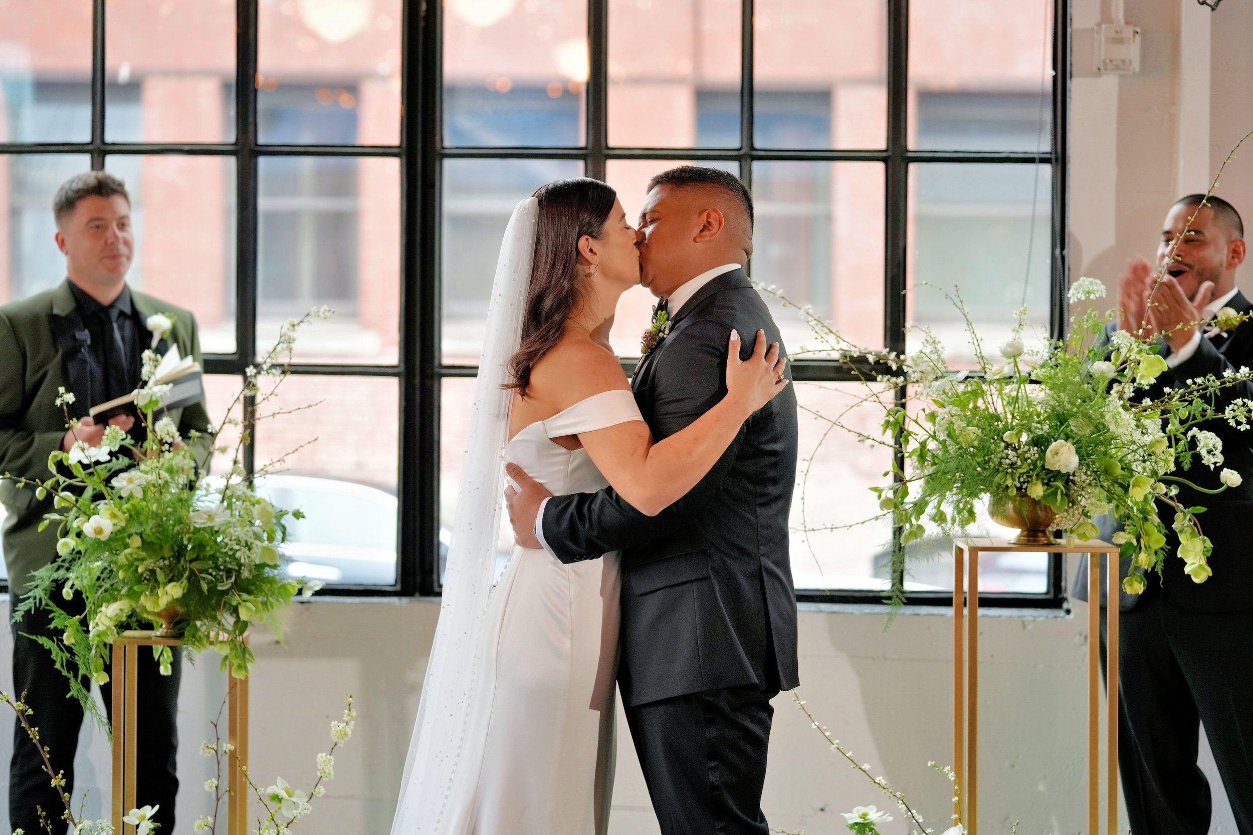 Bride and groom share their first kiss in front of lush white and green altar arrangements at Castaway Portland, designed with local spring blooms by Roots of Joy.