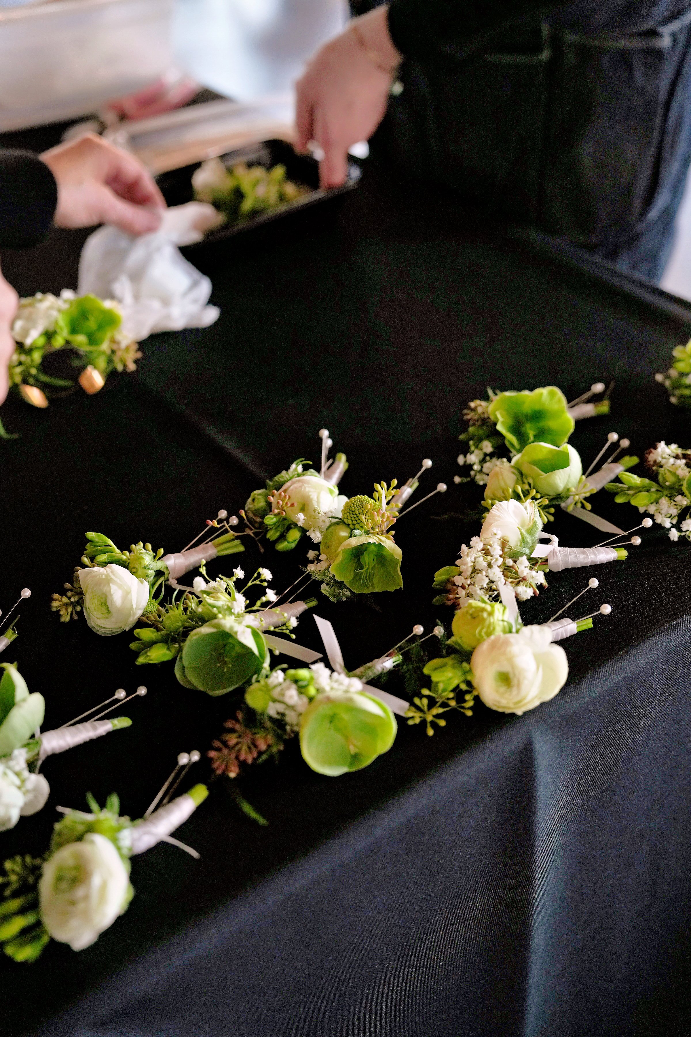 White and green wedding boutonnieres with ranunculus, hellebores, and baby’s breath being arranged on a black linen table during setup for a contemporary Portland wedding.