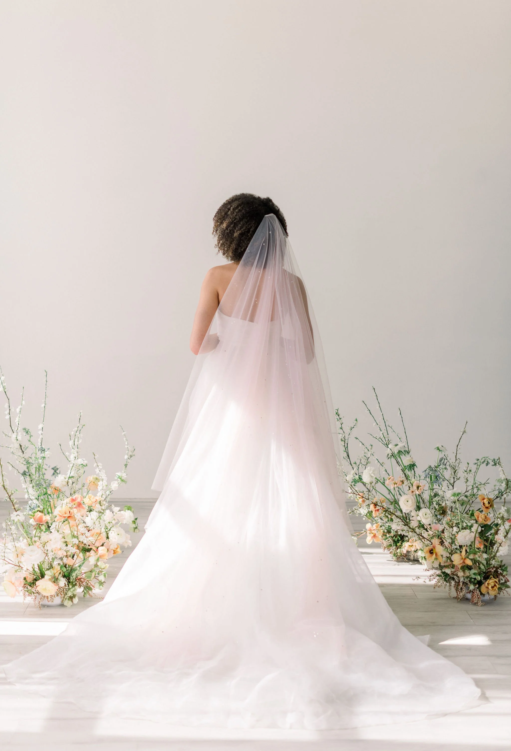 Bride in soft tulle veil by Tessa Kim stands in a minimal studio space, holding a pastel bouquet and surrounded by romantic garden-inspired floral installations by Roots of Joy.