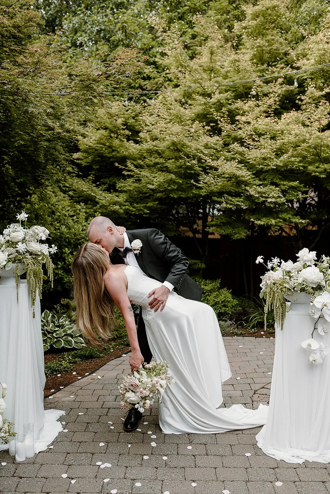 Bride and groom share a romantic dip kiss during their backyard wedding ceremony, framed by modern white floral arrangements by Roots of Joy in Portland, Oregon.