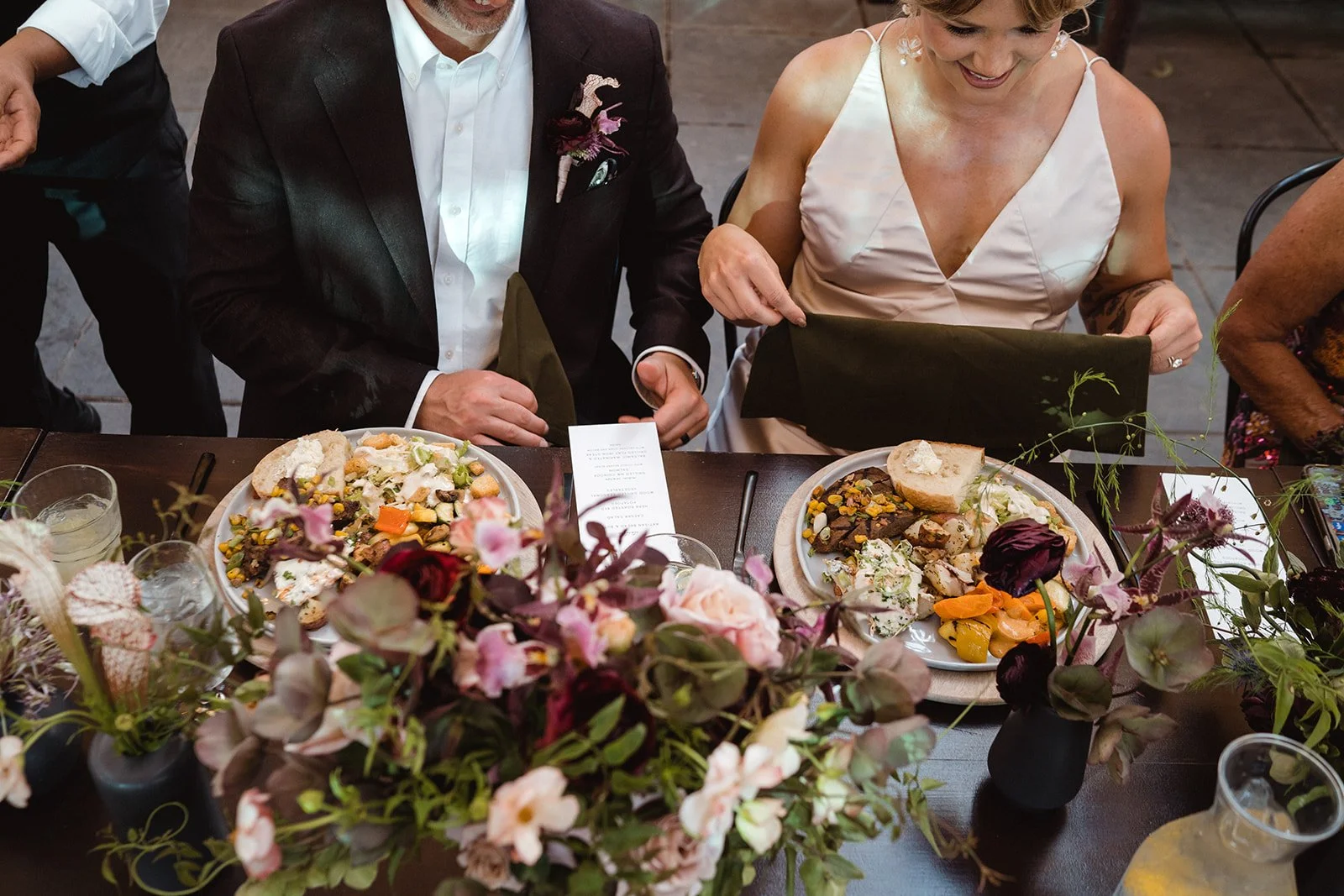Bride and groom seated for dinner surrounded by lush floral arrangements featuring ranunculus, sweet pea, and greenery by Roots of Joy.