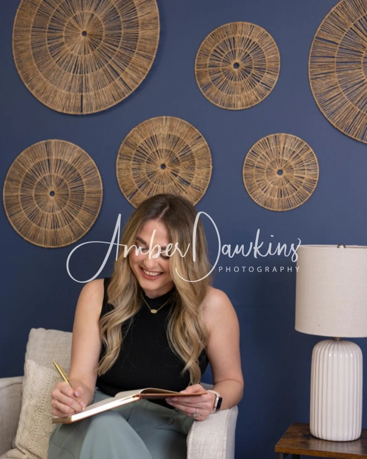 A woman with long wavy blonde hair, wearing a black sleeveless top, sitting on a beige sofa, smiling and writing in a notebook, with a small pillow behind her, a beige table lamp on her right, and a wall decorated with circular woven wall art on a navy blue background.