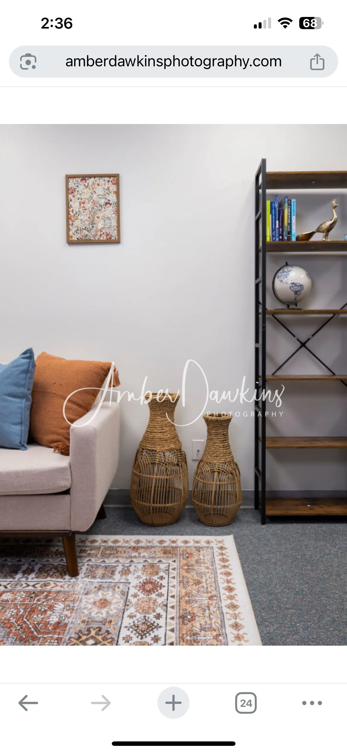 Living room corner with beige sofa, blue and orange pillows, two woven vases, a white bookshelf with decorative items and books, a framed artwork on white wall, and a patterned rug on a gray carpet.