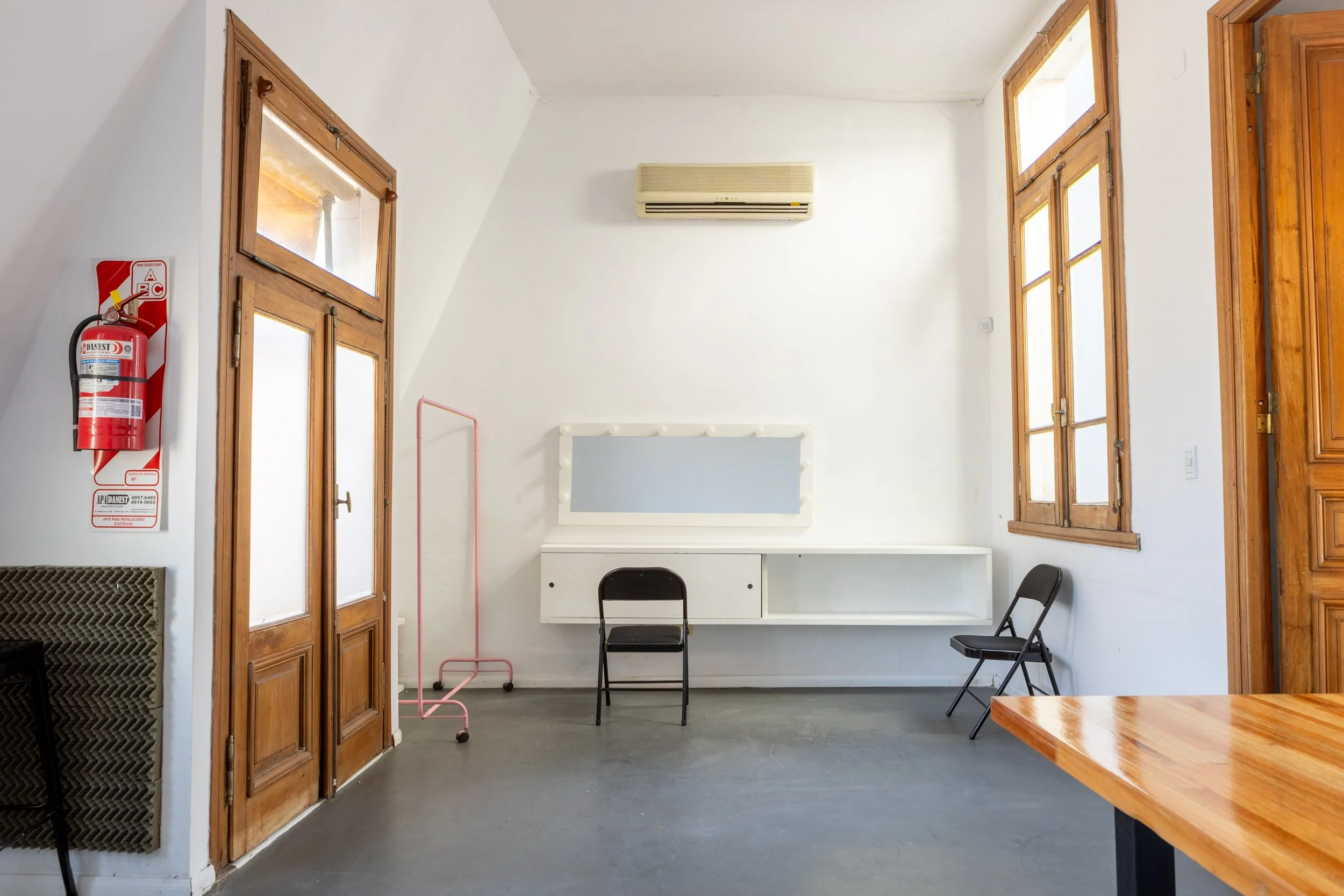 Espacio de oficina moderno con mesas de madera, sillas blancas, lámparas colgantes, un estante naranja con equipos de sonido, y una sala de maquillaje con luz rosa al fondo.