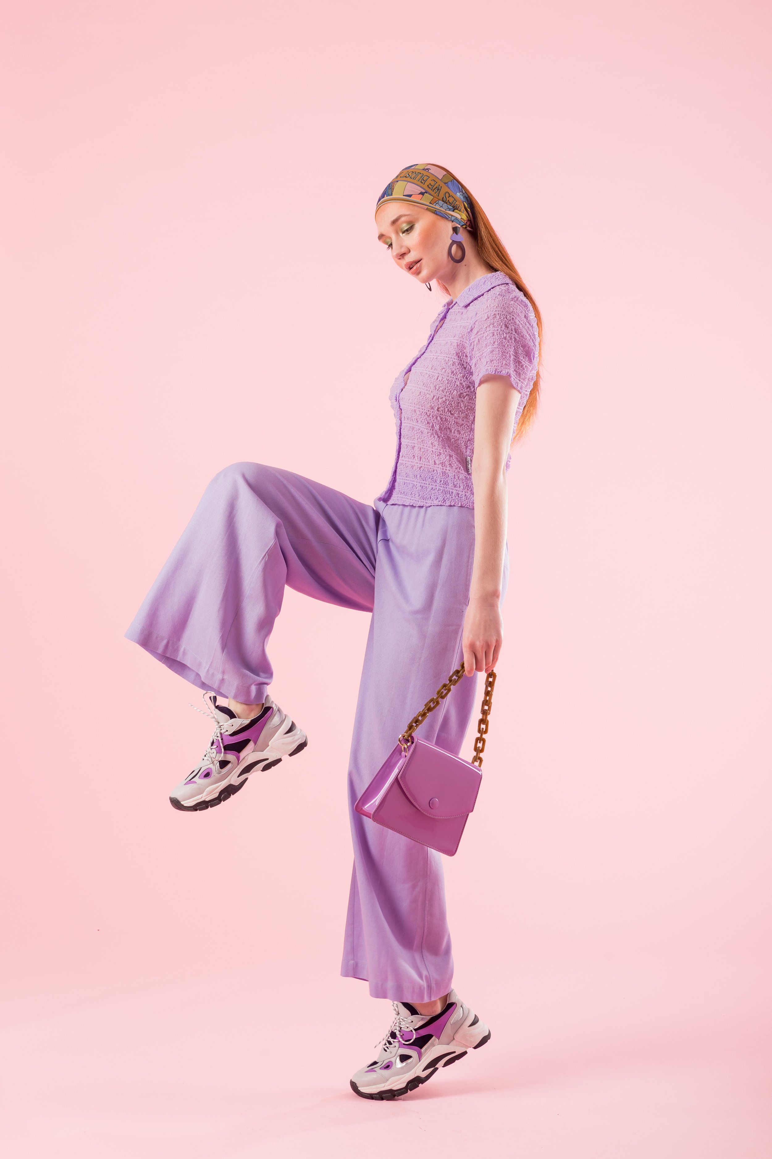 Persona con ropa lila, bufanda en la cabeza, zapatillas deportivas y bolso pequeño, fondo rosa.