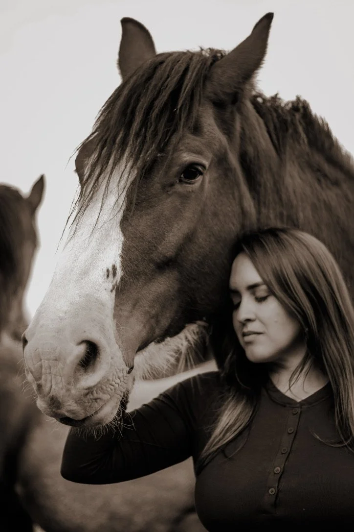 A woman with long hair and a dark shirt gently resting against a large horse with a white blaze on its face, both with eyes closed in a peaceful moment.