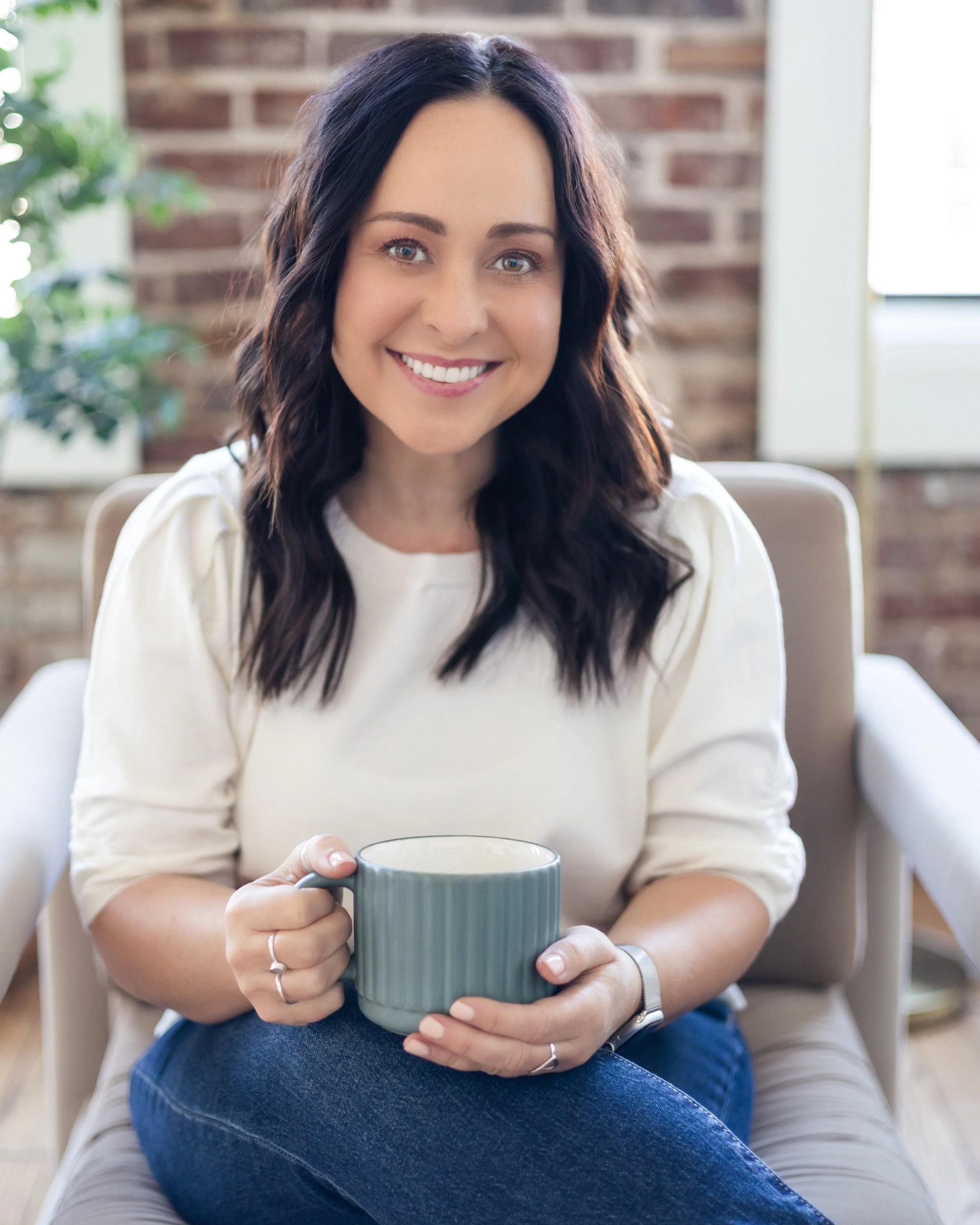 Sara Stockinger sitting comfortably in a chair, smiling warmly while holding a mug, in a cozy and inviting space with a brick wall and natural light.