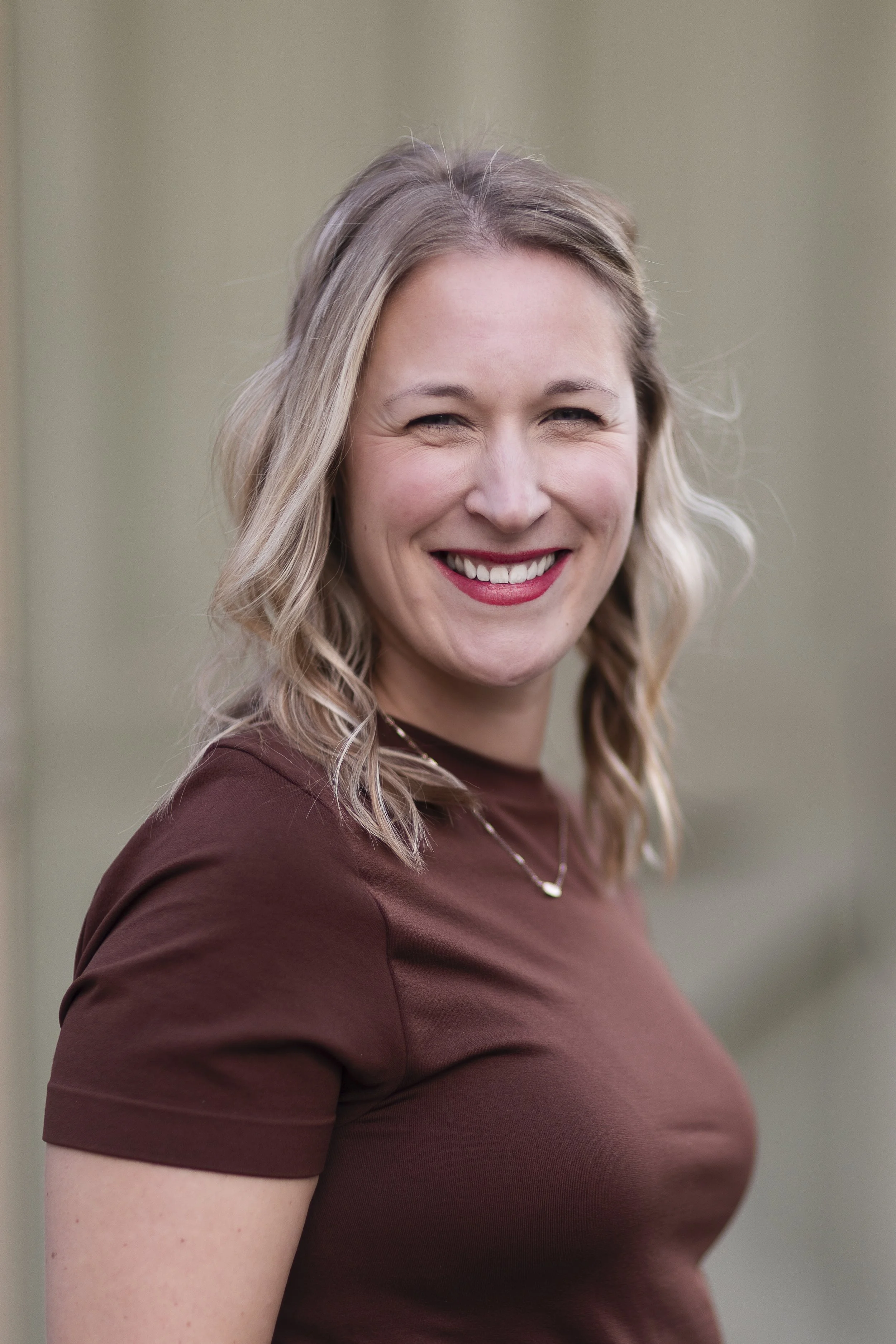 A smiling woman with blonde, wavy hair wearing a brown T-shirt and a delicate necklace, standing outdoors.