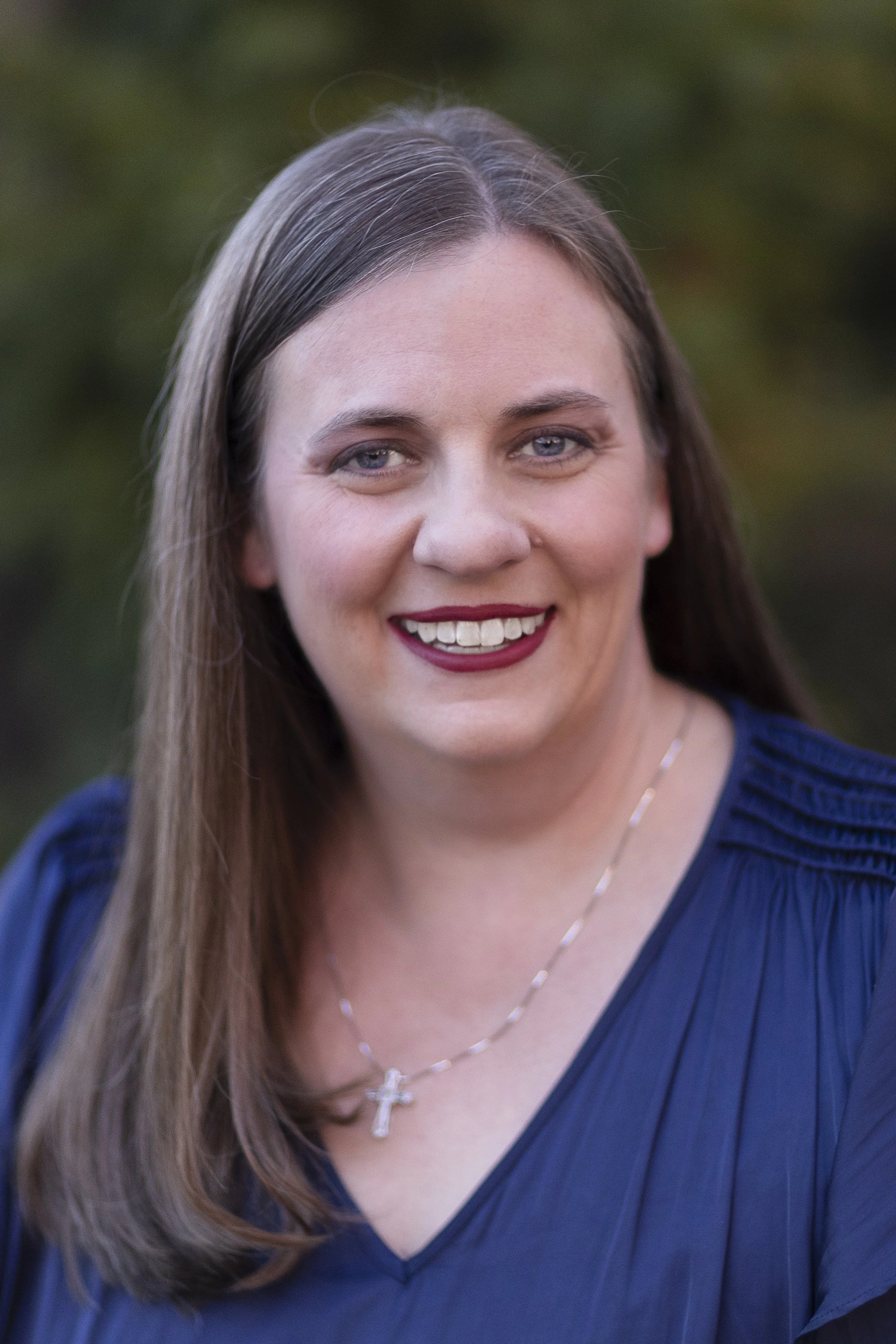 A smiling woman with long brown hair and blue eyes, wearing a navy blue top and a silver cross necklace, outdoors with a blurred green background.
