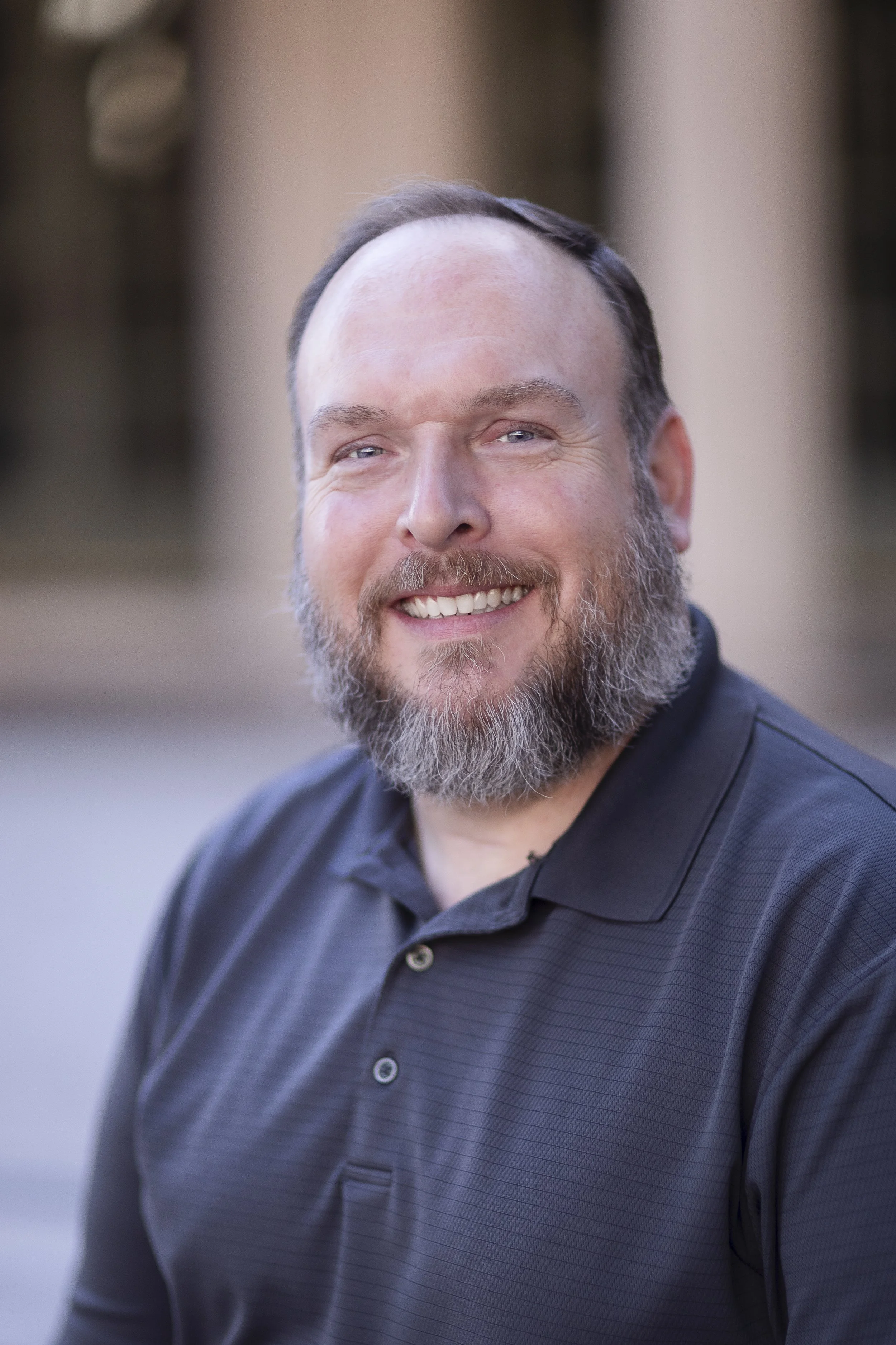 A smiling man with a beard and short hair, wearing a dark polo shirt, standing outdoors with a blurred architectural background.