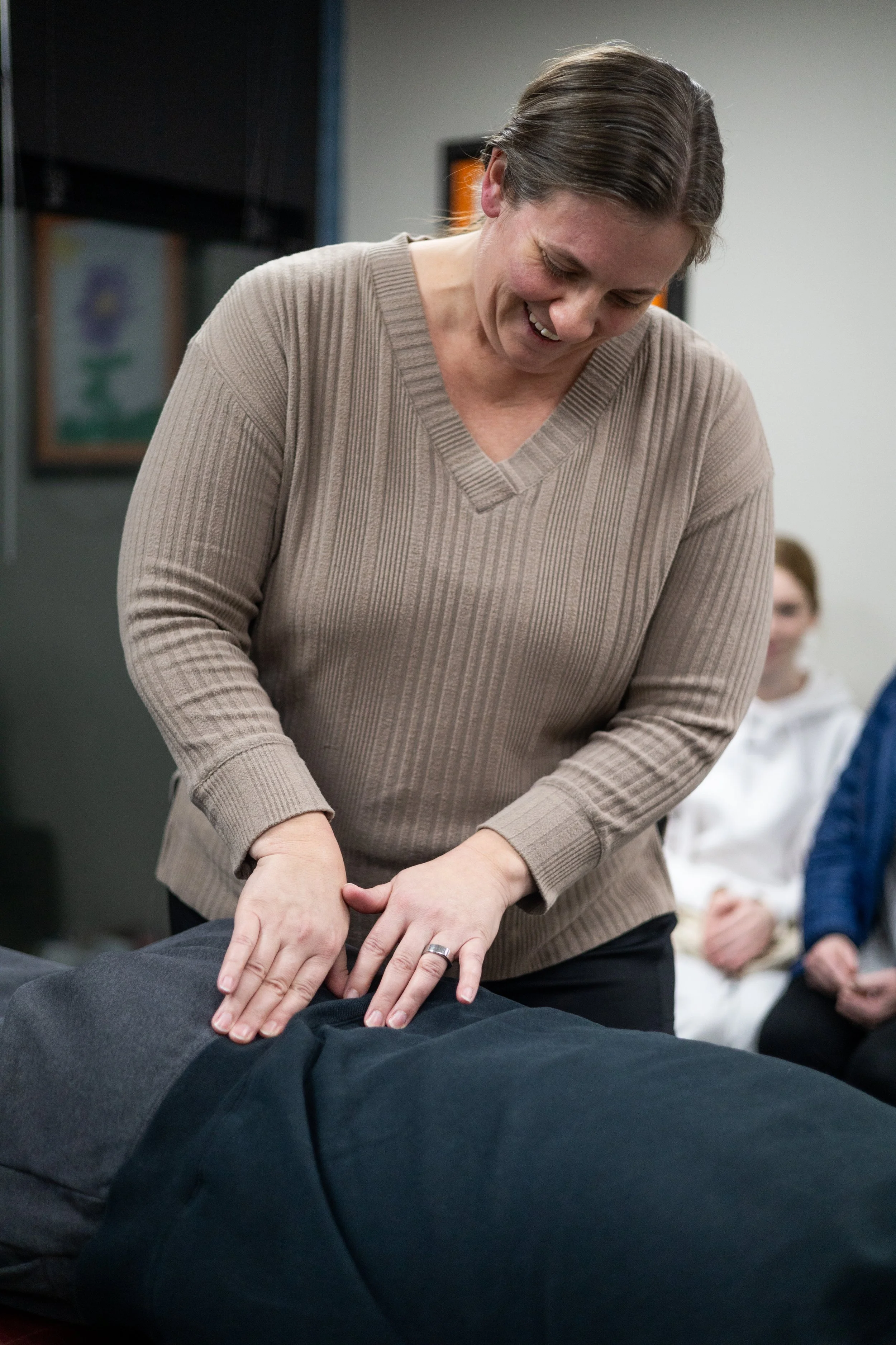 A woman performing chest compressions during CPR training, with a group of people observing in the background.