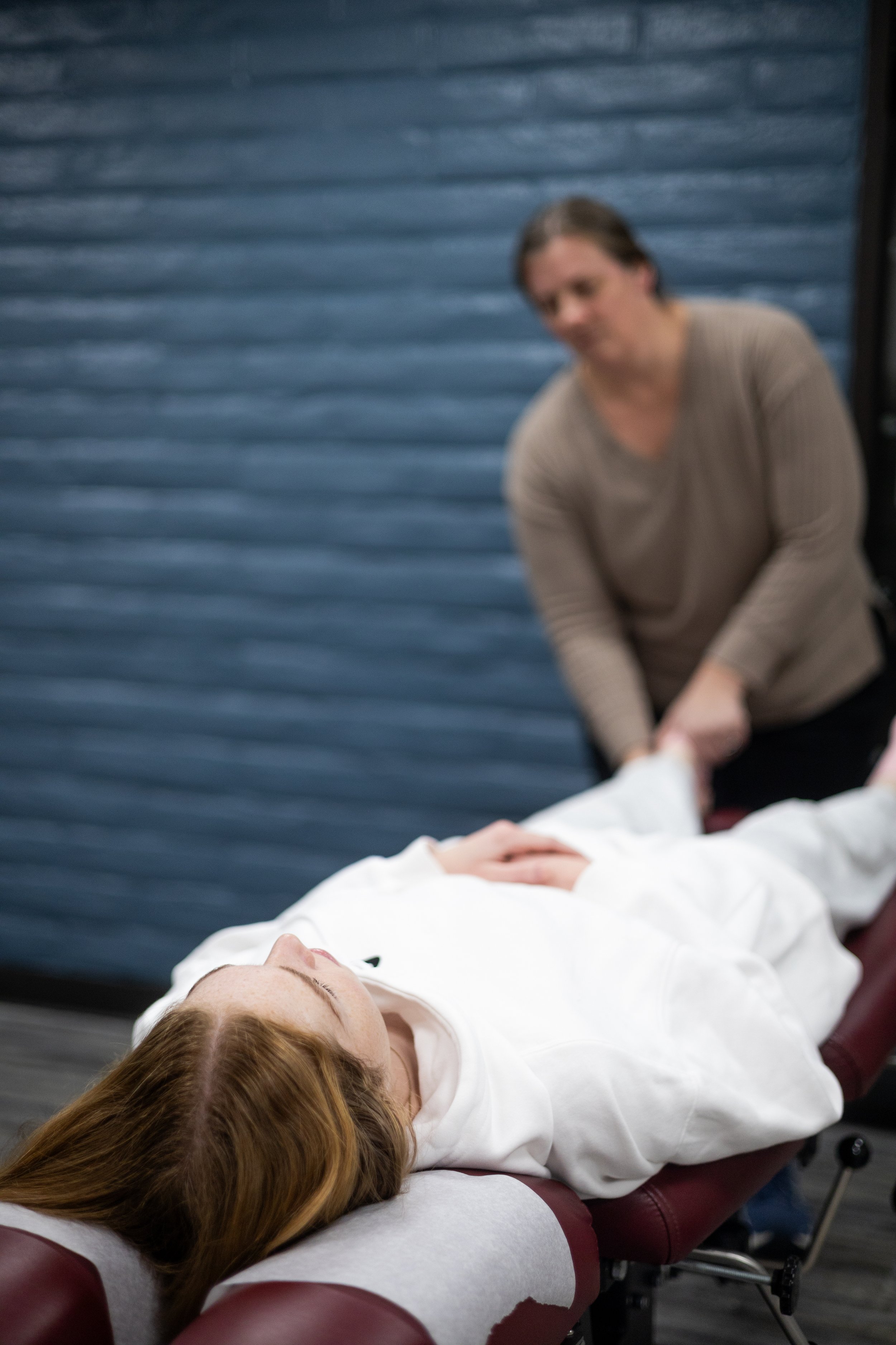 A woman lying on her back on a padded table with her eyes closed, while a person in the background holds her hand, with a blue textured wall behind them.