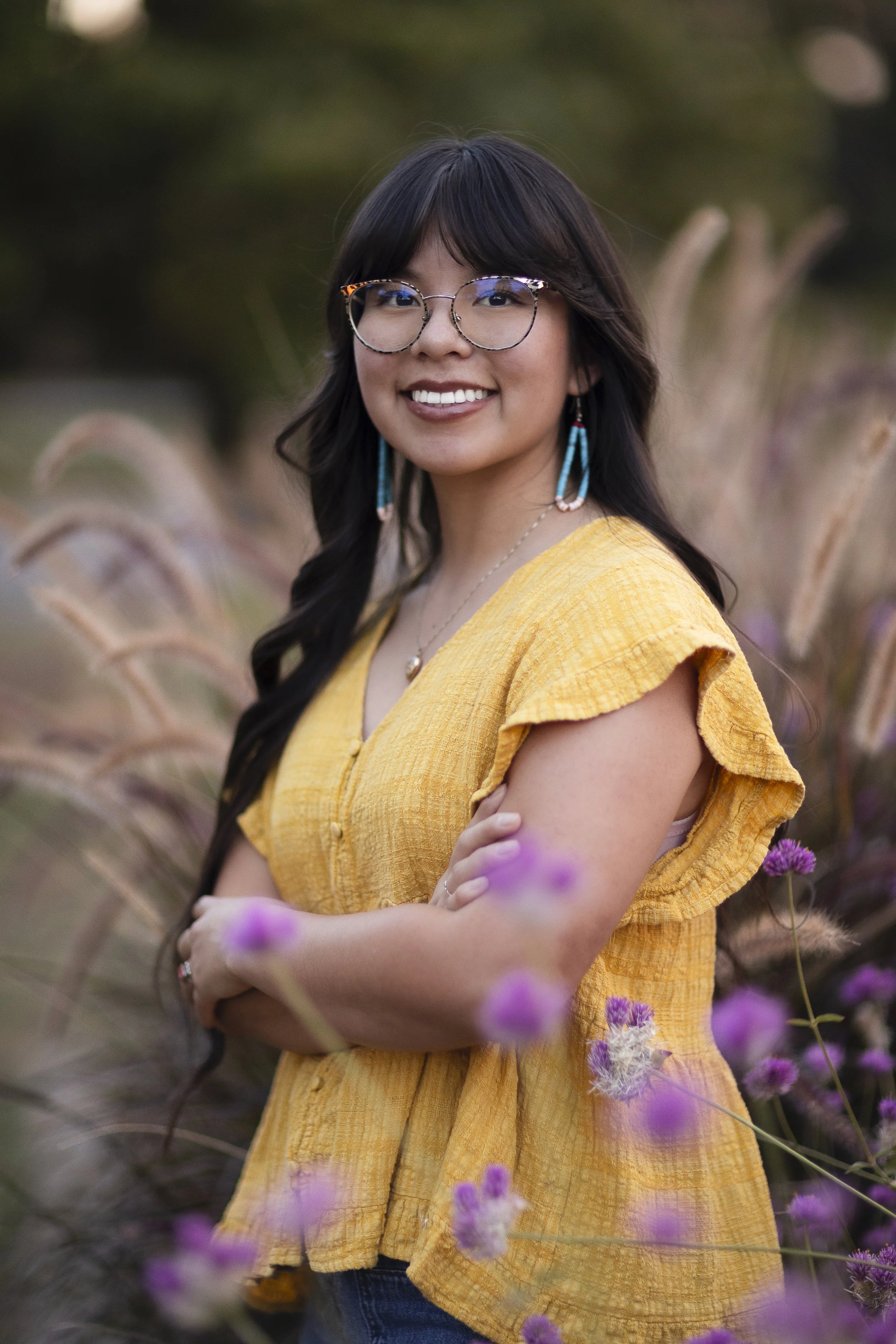 A woman with long dark hair, glasses, and turquoise earrings, wearing a yellow blouse, standing outdoors among purple flowers, smiling at the camera.