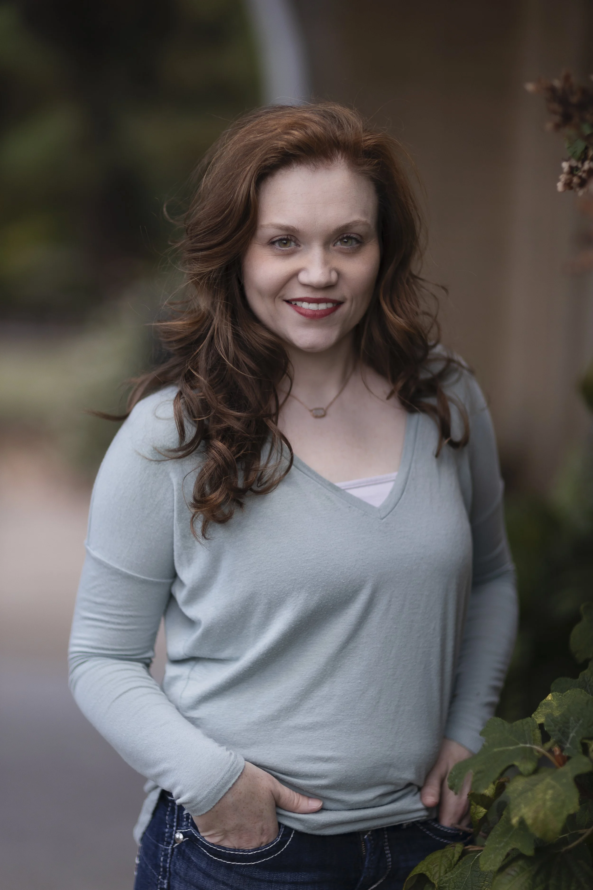 Portrait of a woman with curly red hair, smiling, outdoors with greenery in the background, wearing a light gray long-sleeve shirt and dark jeans.