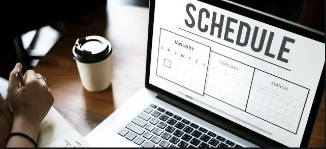 Laptop on a wooden desk displaying a digital calendar with the heading "Schedule." Nearby, a person's hand holding a pen and a coffee cup.