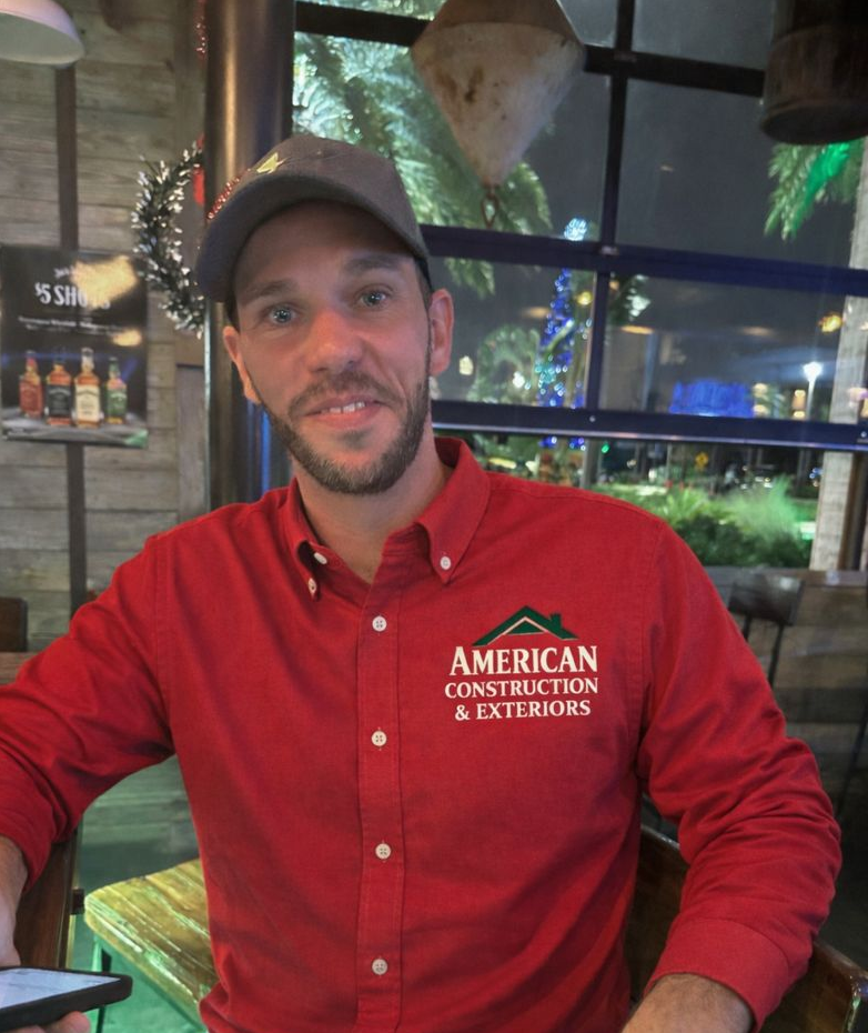 Man wearing red shirt with 'American Construction & Exteriors' logo, sitting indoors at a restaurant.