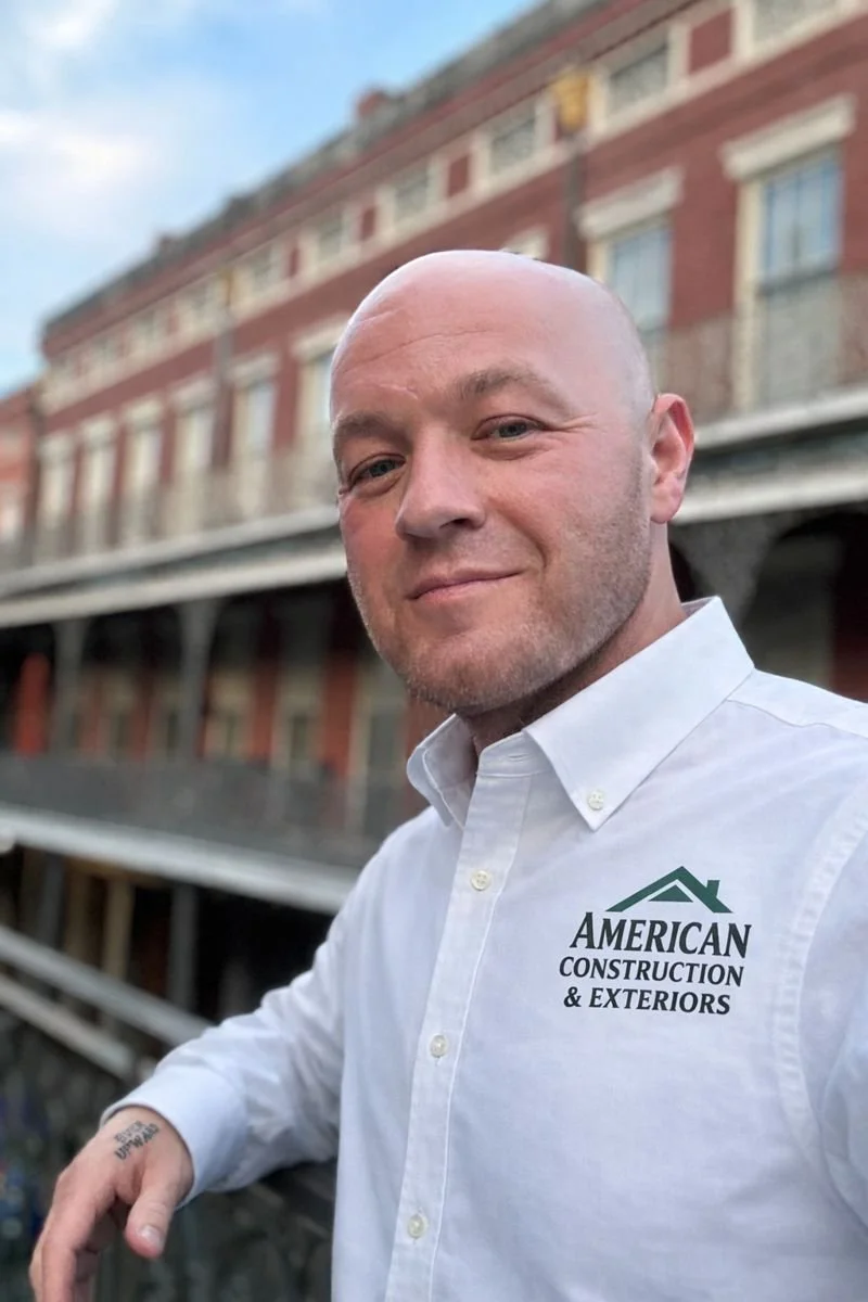 A man in a white button-up shirt with the logo of American Construction & Exteriors poses outdoors in front of a multi-story brick building with ornate windows and balconies, during daytime.