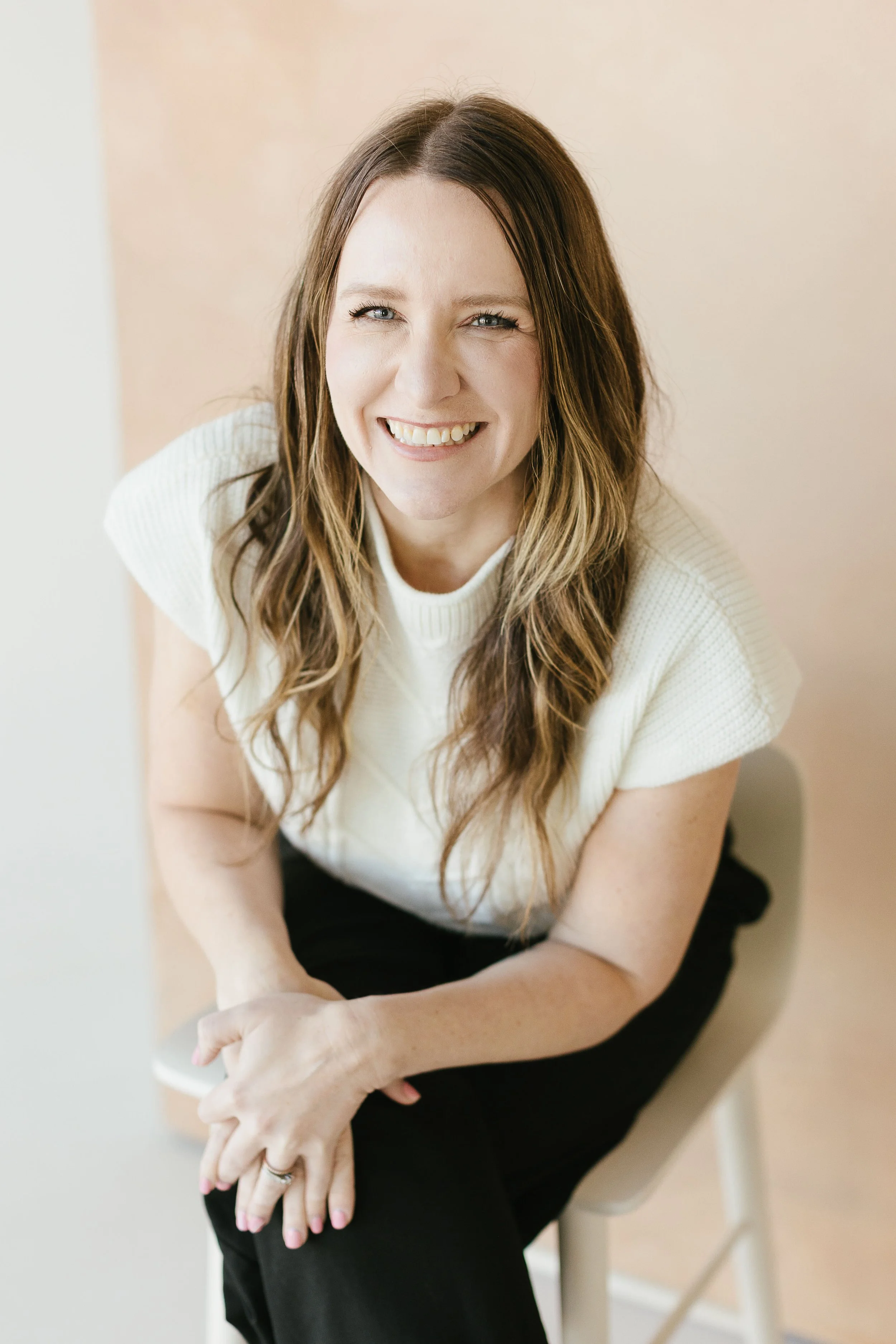 A woman with long brown hair and blue eyes, wearing a sleeveless black top and a stethoscope around her neck, sitting in front of green plants with large leaves.