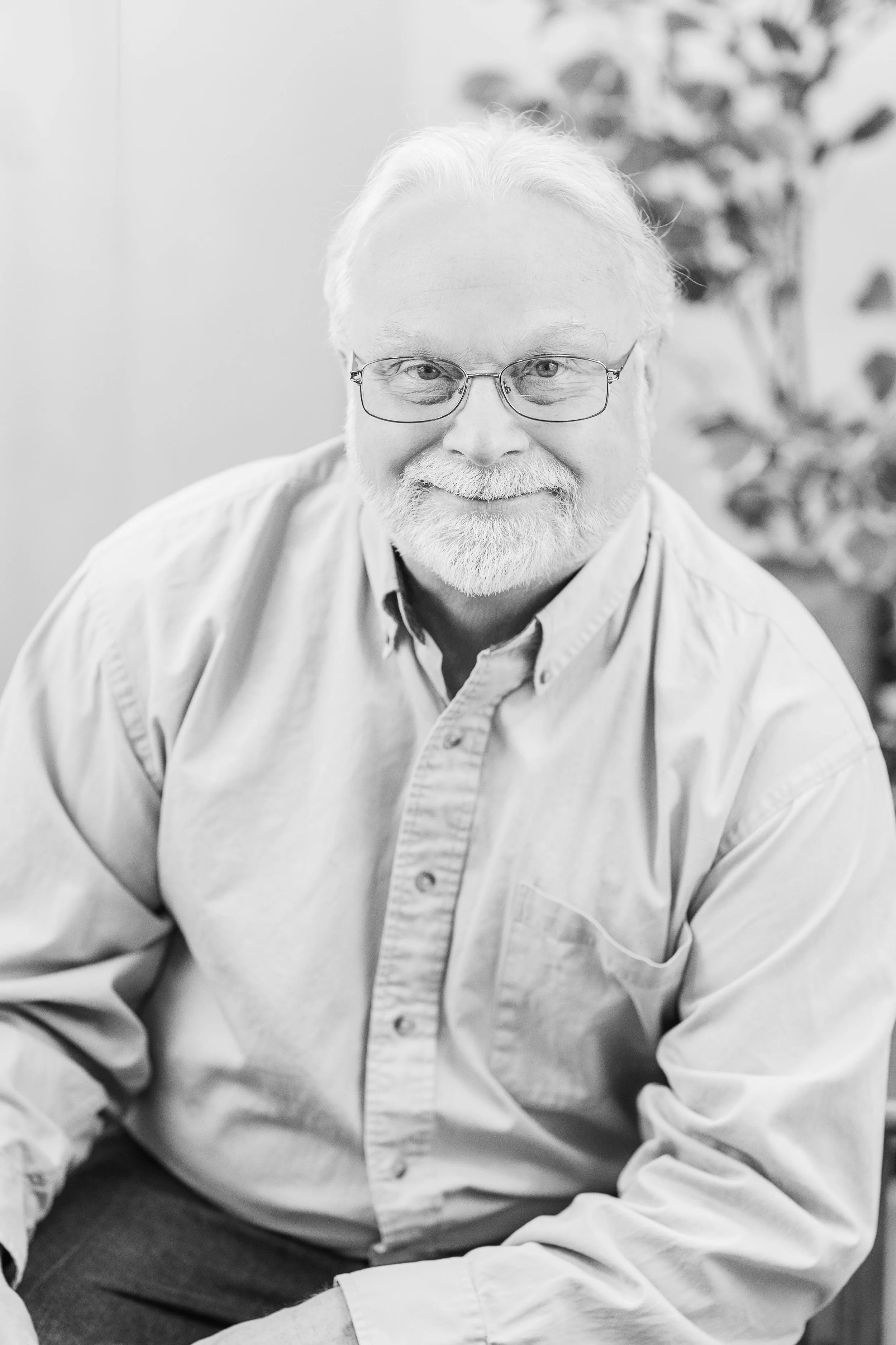 A smiling elderly man with glasses, white hair, and a beard, wearing a light-colored button-up shirt, sitting indoors with a blurred plant in the background.