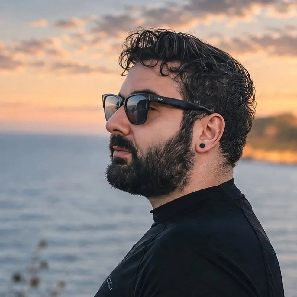 Profile of a man with dark hair and beard, wearing sunglasses and a black shirt, looking at the water during sunset.