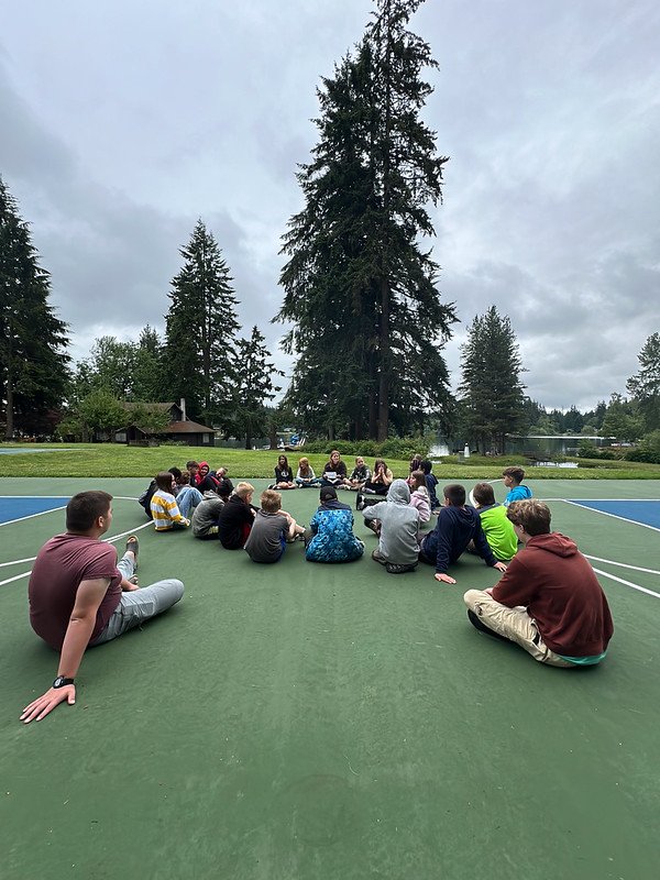 Group of people sitting in a circle on a sports court with a backdrop of tall trees and a cloudy sky.