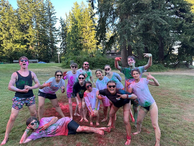 Group of people having a colorful outdoor celebration on a grassy field with trees in the background, wearing casual clothes and covered in colorful powder.