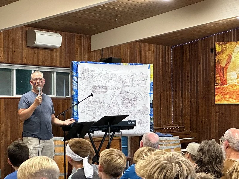 Man speaking with microphone in front of a map-like illustration, addressing an audience in a wooden room.