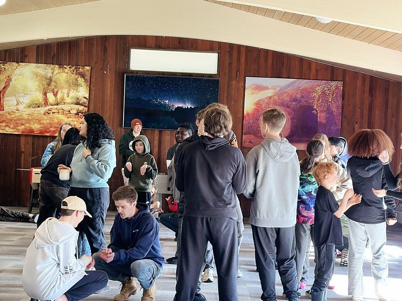 A group of people in casual attire gathered indoors, engaged in conversation and activities. The room has wooden paneling and three colorful landscape paintings on the wall.