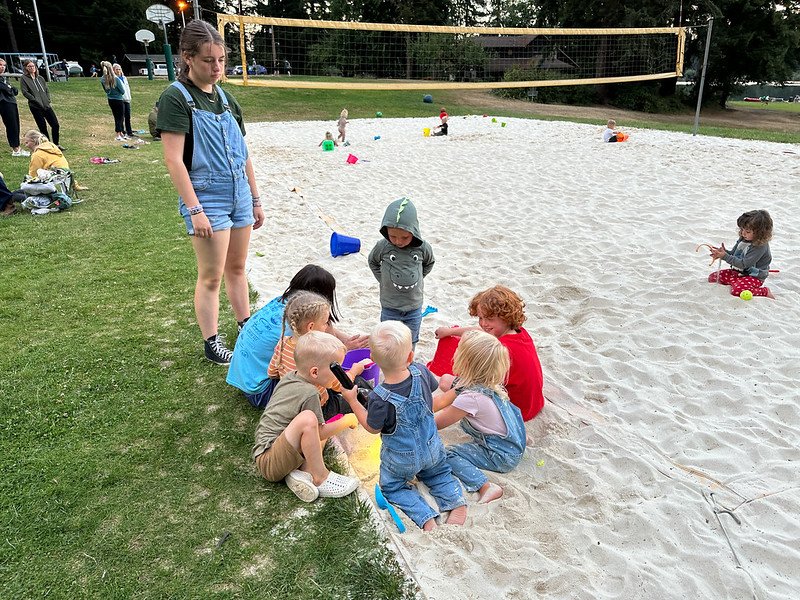 Children playing in a sand area with toys, supervised by an adult, near a volleyball net at a park.