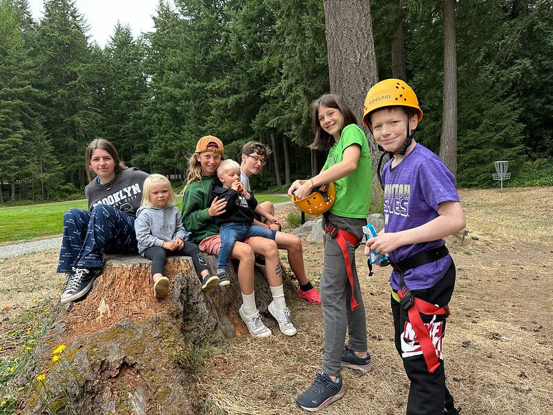Group of kids in outdoor setting with climbing gear, sitting on tree stump and standing nearby in forested area.