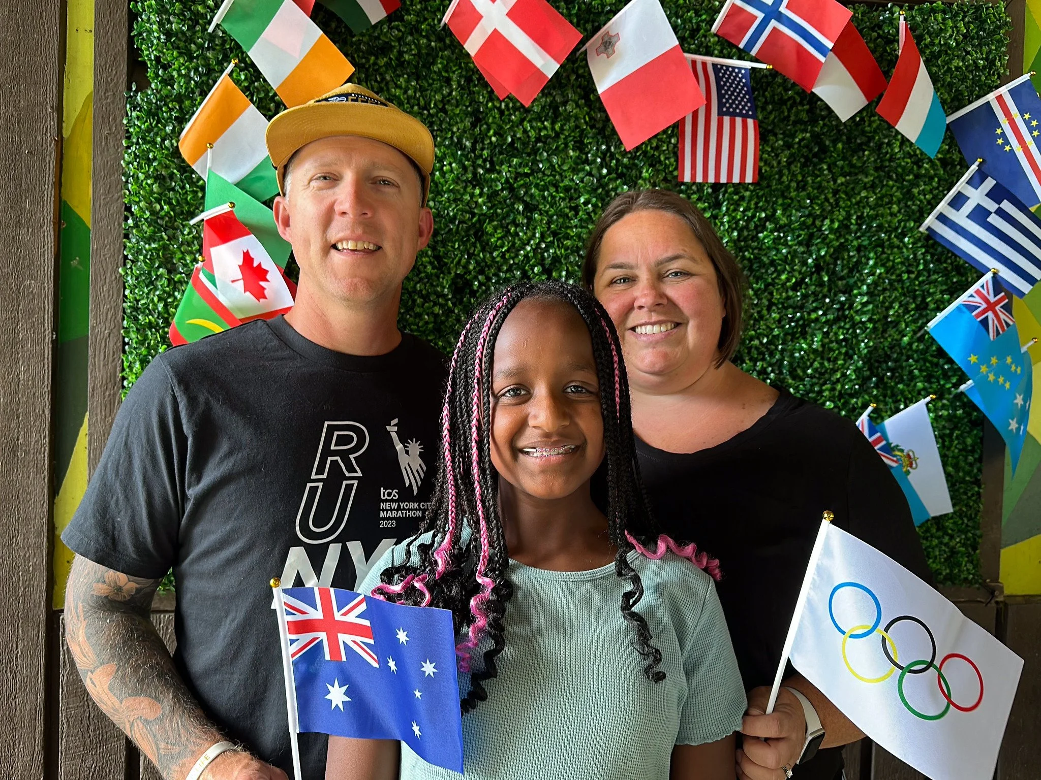 A group of three people smiling for a photo, holding flags of Australia and the Olympic rings, standing in front of a backdrop decorated with small international flags and greenery.