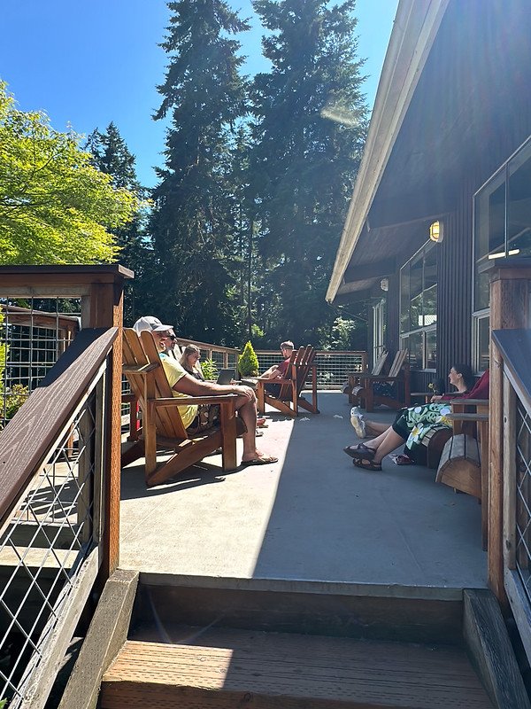 People sitting on a wooden deck in Adirondack chairs, surrounded by trees on a sunny day.
