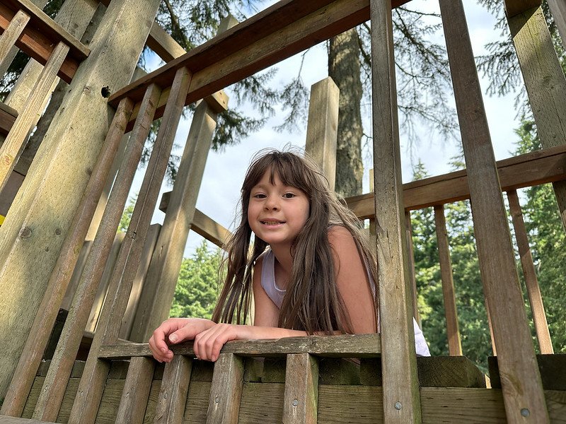 A young girl smiling and leaning out from a wooden playground structure surrounded by trees.