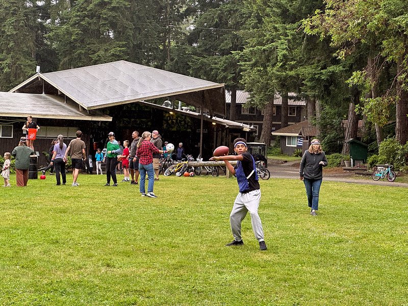 Group of people playing football on a grassy field near a pavilion and surrounded by trees.