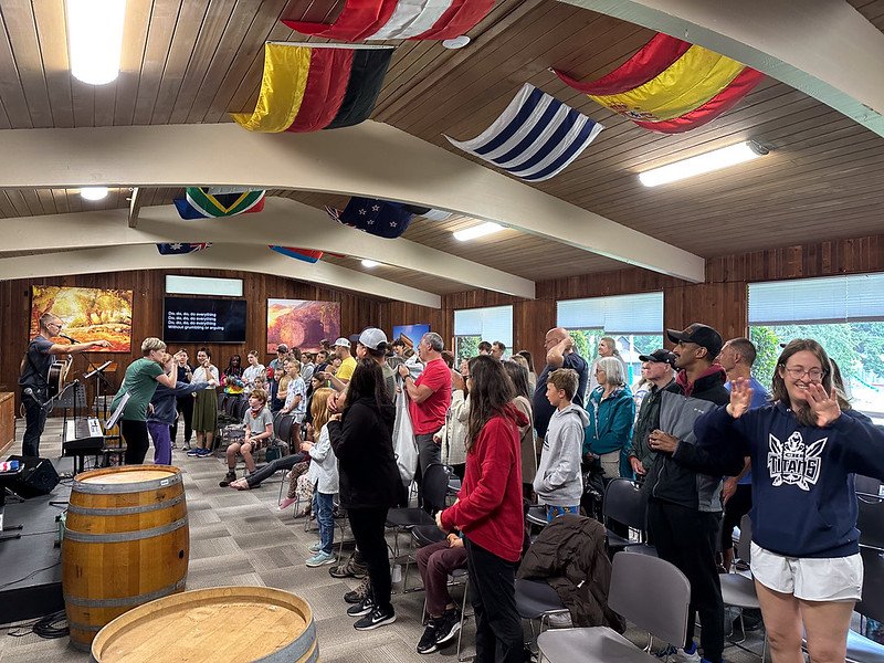 Group of people gathered in a hall with wooden walls, listening to a speaker. Flags hang from the ceiling. Attendees include men, women, and children, some standing and raising hands.