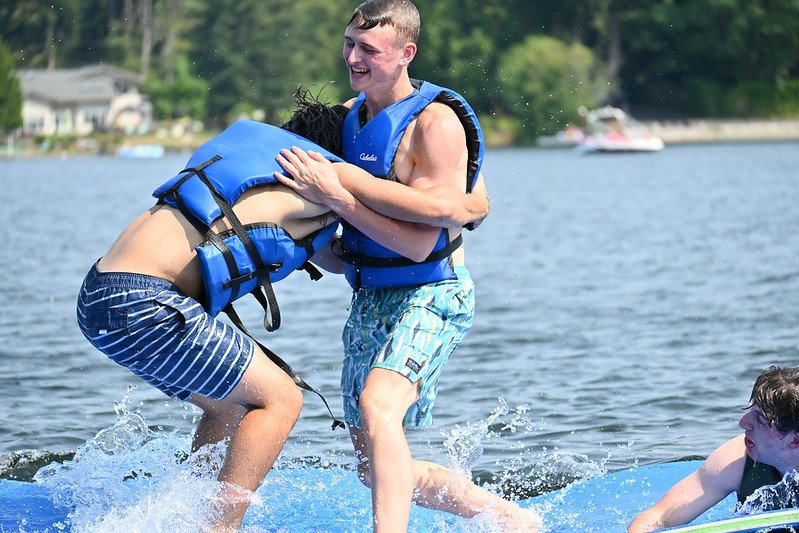 Two people in blue life jackets playfully wrestling on a floating mat in a lake.