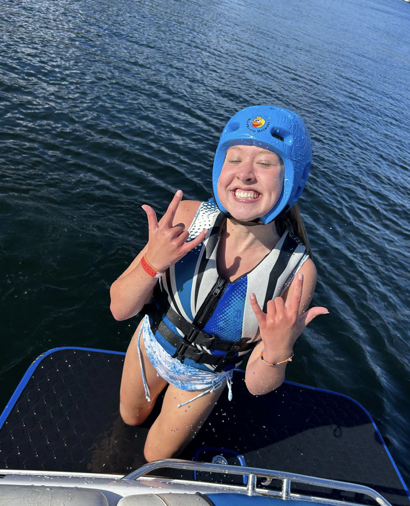 Young girl on a boat wearing a blue helmet, smiling, making a rock and roll hand sign, water in the background.