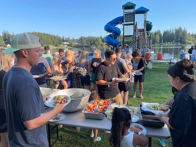 People enjoying a picnic buffet with food trays and a water slide at a park by a lake.