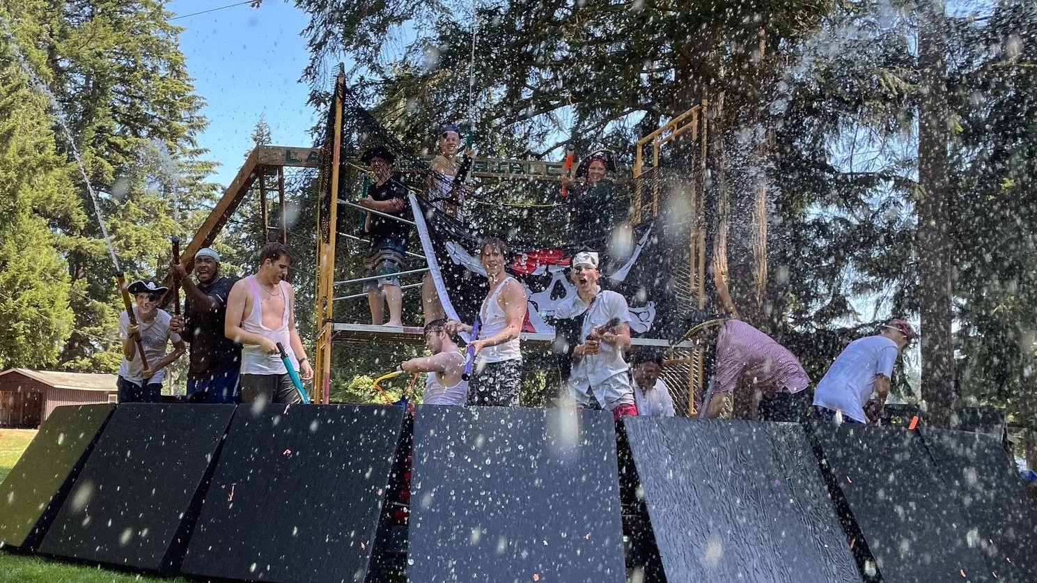 People celebrating with water on a wooden pirate ship play structure outdoors, wearing costumes and rain gear, surrounded by trees and blue sky.