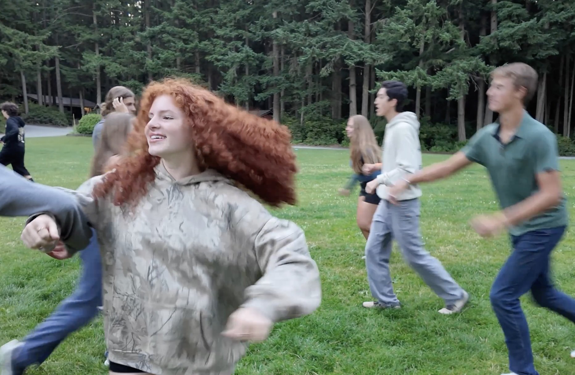 Group of young people, including a red-haired girl, running and playing on a grassy field with trees in the background.