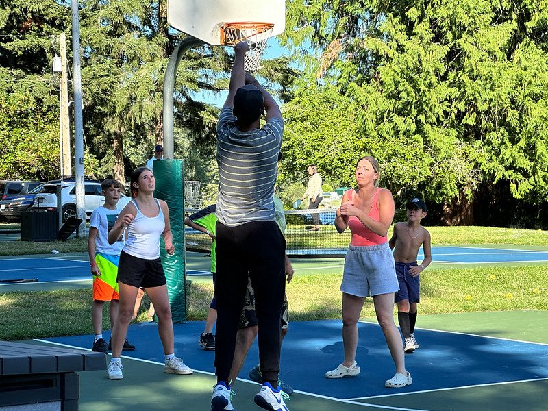 Group of people playing basketball on an outdoor court with trees and grass nearby.